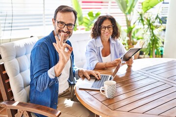 Middle age couple relaxing at home terrace doing ok sign with fingers, smiling friendly gesturing...