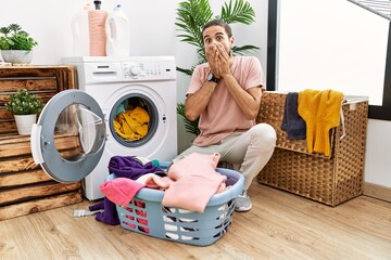 Young hispanic man putting dirty laundry into washing machine shocked covering mouth with hands for mistake. secret concept.