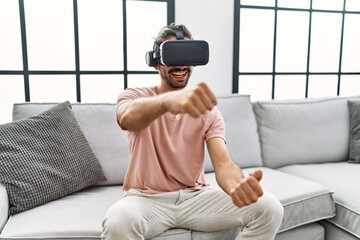 Handsome hispanic man wearing virtual reality glasses driving on videogame at the living room at home