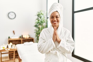 Young brunette woman wearing towel and bathrobe standing at beauty center praying with hands together asking for forgiveness smiling confident.