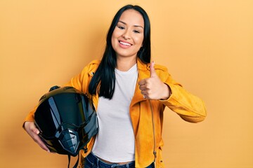 Beautiful hispanic woman with nose piercing holding motorcycle helmet smiling happy and positive, thumb up doing excellent and approval sign