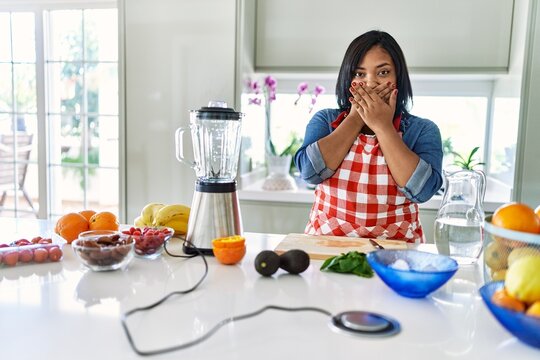 Young hispanic woman making healthy smoothie shocked covering mouth with hands for mistake. secret concept.