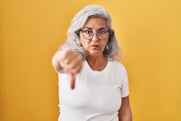 Middle age woman with grey hair standing over yellow background looking unhappy and angry showing...