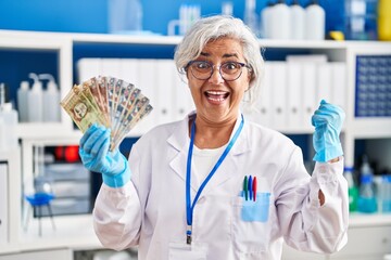 Middle age woman with grey hair working at scientist laboratory holding polish zloty banknotes screaming proud, celebrating victory and success very excited with raised arms