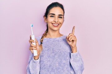 Young hispanic girl holding electric toothbrush smiling with an idea or question pointing finger...