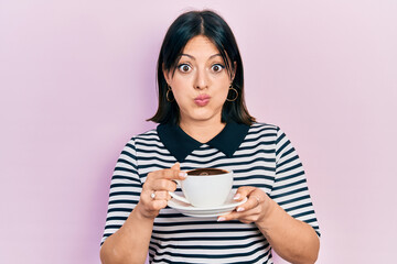 Young hispanic woman drinking a cup of coffee puffing cheeks with funny face. mouth inflated with...