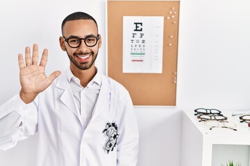 Fototapeta na wymiar African american optician man standing by eyesight test showing and pointing up with fingers number five while smiling confident and happy.