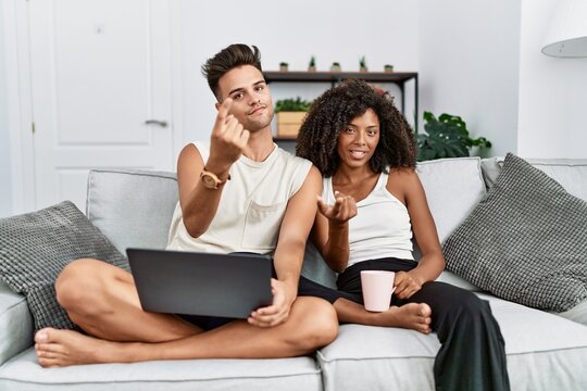 Young interracial couple using laptop at home sitting on the sofa beckoning come here gesture with hand inviting welcoming happy and smiling