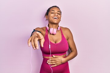 Young african american girl wearing gym clothes and using headphones laughing at you, pointing...
