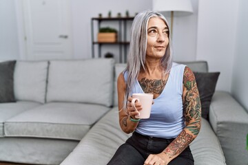 Middle age grey-haired woman drinking coffee sitting on the sofa at home looking away to side with smile on face, natural expression. laughing confident.
