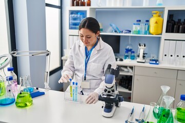 Young hispanic woman wearing scientist uniform holding test tubes at laboratory