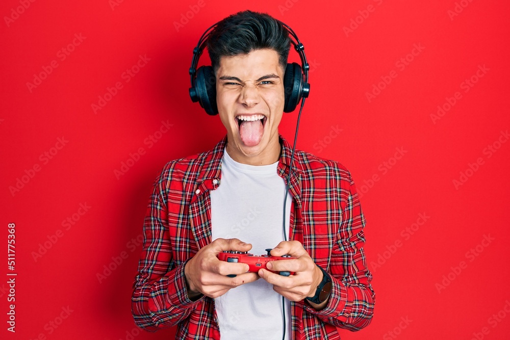 Wall mural young hispanic man playing video game holding controller sticking tongue out happy with funny expres