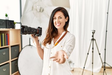Beautiful caucasian woman working as photographer at photography studio smiling cheerful offering palm hand giving assistance and acceptance.