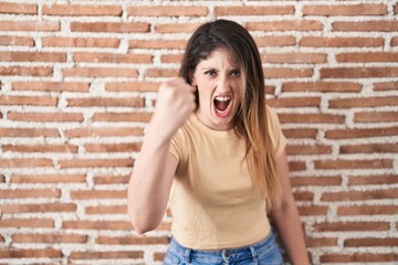 Young brunette woman standing over bricks wall angry and mad raising fist frustrated and furious...