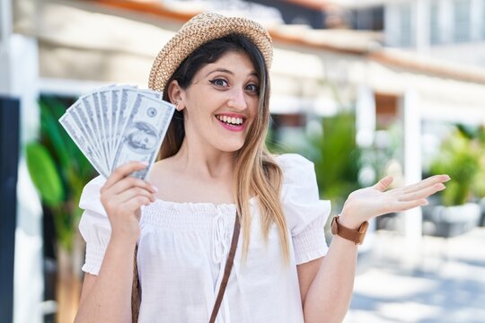 Young brunette woman holding dollars banknotes celebrating achievement with happy smile and winner expression with raised hand