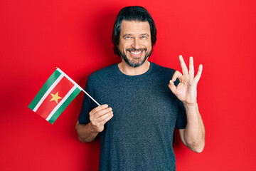 Middle age caucasian man holding suriname flag doing ok sign with fingers, smiling friendly...
