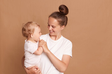 Indoor shot of mother playing with baby girl, happy family having fun at home, cheerful sweet kid in mom's arms, mom and child isolated over brown background.