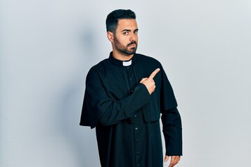 Handsome hispanic man with beard wearing catholic priest robe pointing with hand finger to the side showing advertisement, serious and calm face