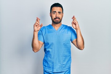 Fototapeta premium Handsome hispanic man with beard wearing blue male nurse uniform gesturing finger crossed smiling with hope and eyes closed. luck and superstitious concept.