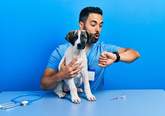 Handsome hispanic veterinary man with beard checking dog health looking at the watch time worried,...