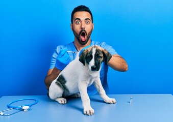 Handsome hispanic veterinary man with beard putting vaccine to puppy dog afraid and shocked with surprise and amazed expression, fear and excited face.