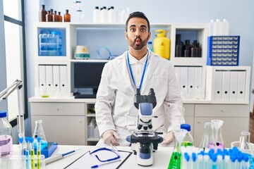 Fototapeta premium Young hispanic man with beard working at scientist laboratory making fish face with lips, crazy and comical gesture. funny expression.