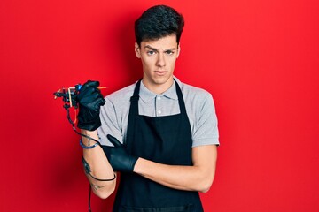 Young hispanic man tattoo artist wearing professional uniform and gloves holding tattooer machine smiling happy and positive, thumb up doing excellent and approval sign