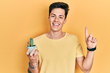 Young hispanic man holding small cactus pot smiling with an idea or question pointing finger with happy face, number one - obrazy, fototapety, plakaty