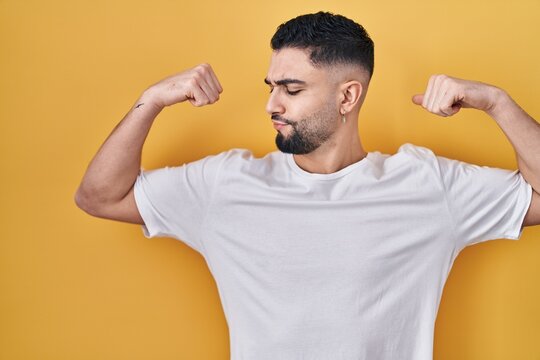 Young handsome man wearing casual t shirt over yellow background showing arms muscles smiling proud. fitness concept.