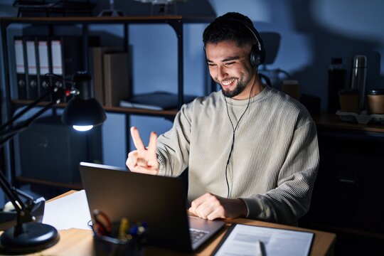 Young handsome man working using computer laptop at night smiling looking to the camera showing fingers doing victory sign. number two.