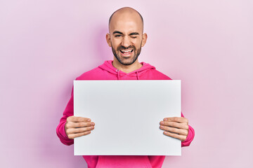 Young bald man holding blank empty banner winking looking at the camera with sexy expression,...