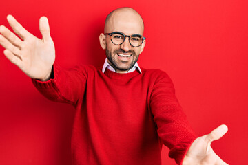 Naklejka premium Young bald man wearing casual clothes and glasses looking at the camera smiling with open arms for hug. cheerful expression embracing happiness.