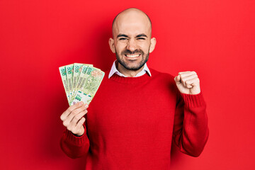 Young bald man holding argentine pesos banknotes screaming proud, celebrating victory and success...