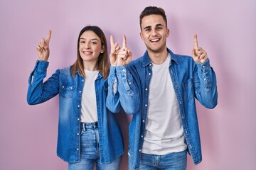 Obraz premium Young hispanic couple standing over pink background smiling amazed and surprised and pointing up with fingers and raised arms.