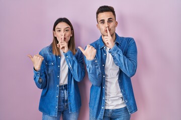 Young hispanic couple standing over pink background asking to be quiet with finger on lips pointing with hand to the side. silence and secret concept.