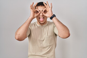 Young arab man wearing casual t shirt doing ok gesture like binoculars sticking tongue out, eyes looking through fingers. crazy expression.