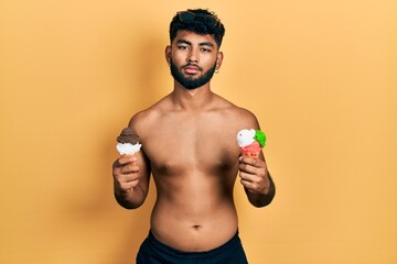 Fototapeta premium Arab man with beard wearing swimwear eating two ice cream cones relaxed with serious expression on face. simple and natural looking at the camera.