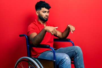 Arab man with beard sitting on wheelchair pointing down looking sad and upset, indicating direction with fingers, unhappy and depressed.