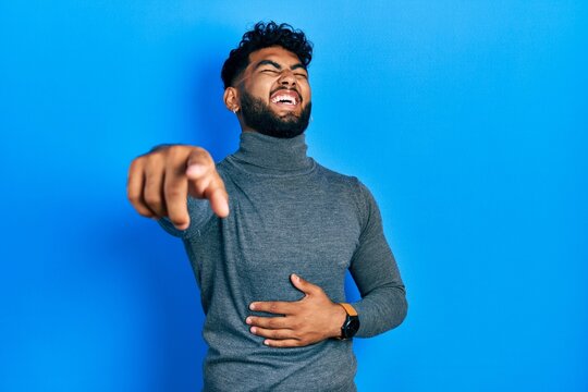 Arab man with beard wearing turtleneck sweater laughing at you, pointing finger to the camera with hand over body, shame expression