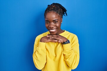 Beautiful black woman standing over blue background laughing nervous and excited with hands on chin...