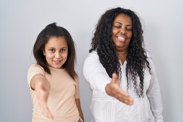 Mother and young daughter standing over white background smiling friendly offering handshake as greeting and welcoming. successful business.