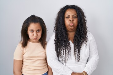 Mother and young daughter standing over white background puffing cheeks with funny face. mouth inflated with air, crazy expression.