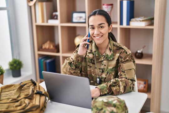 Young hispanic woman army soldier using laptop and talking on martphone at home
