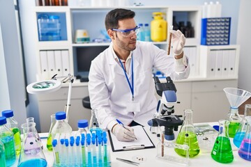 Young hispanic man scientist holding test tube writing report at laboratory