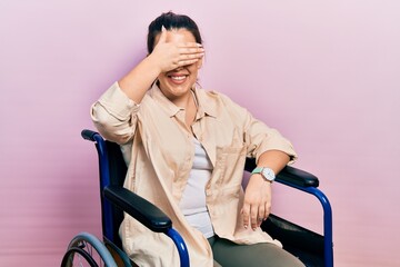 Young hispanic woman sitting on wheelchair smiling and laughing with hand on face covering eyes for surprise. blind concept.