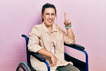 Young hispanic woman sitting on wheelchair pointing finger up with successful idea. exited and happy. number one.