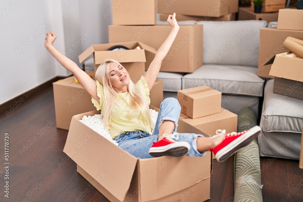 Wall mural young blonde woman smiling confident sitting in cardboard box at new home