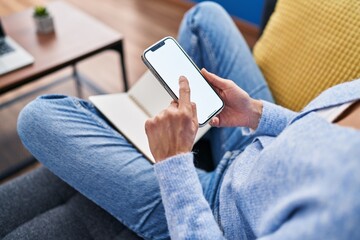 Young woman using smartphone and reading book sitting on sofa at home