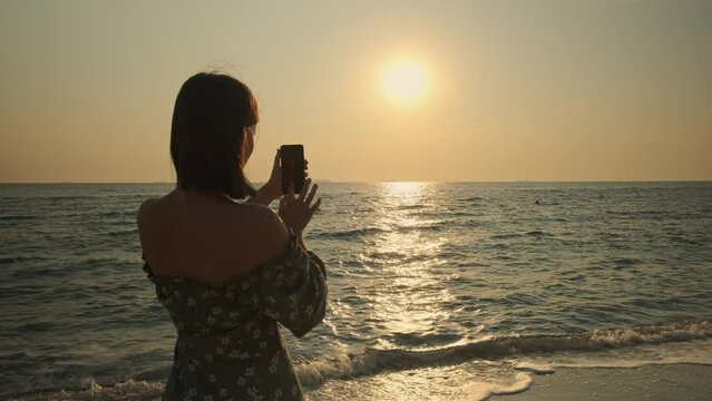 Woman takes photo at the beach. Female tourist taking picture on the phone of the morning sun by the sea. Travel, recreation concept.