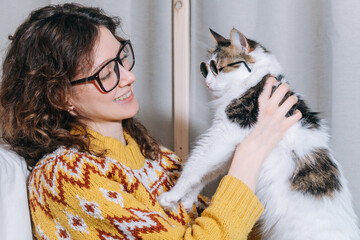 Portrait of a young woman in glasses with curly hair holding a white cat in small glasses sitting on the floor by the bed at home.
